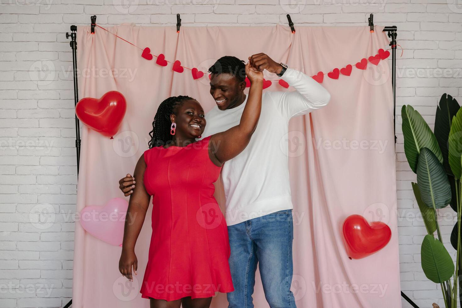 Smiling young African couple dancing on pink fabric background with heart shaped balloons photo