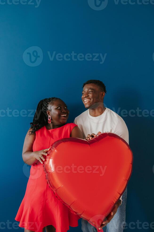 Happy young African couple holding red heart shaped balloon while celebrating Valentines day on blue background photo
