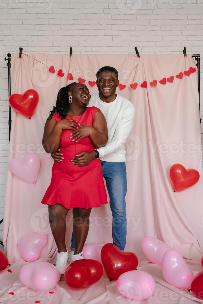 Full length of happy young African couple embracing on pink background with heart shaped balloons photo