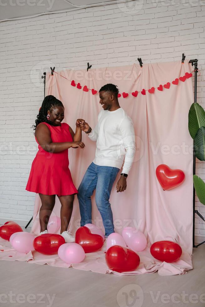Full length of romantic young African couple dancing on pink fabric background with heart shaped balloons photo