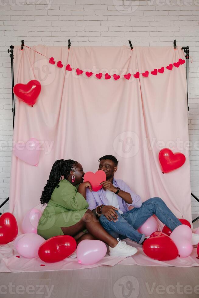 Romantic African couple covering faces with heart shaped Valentine card while sitting on pink fabric background photo