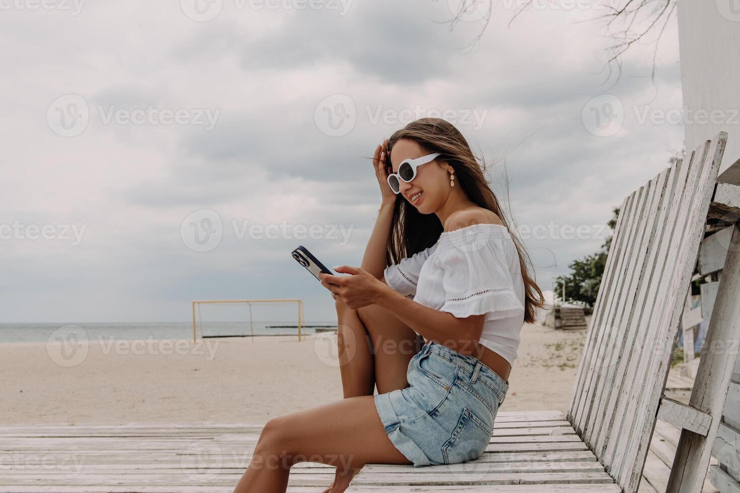 Happy young woman using smart phone while sitting on the wooden pallet seaside photo