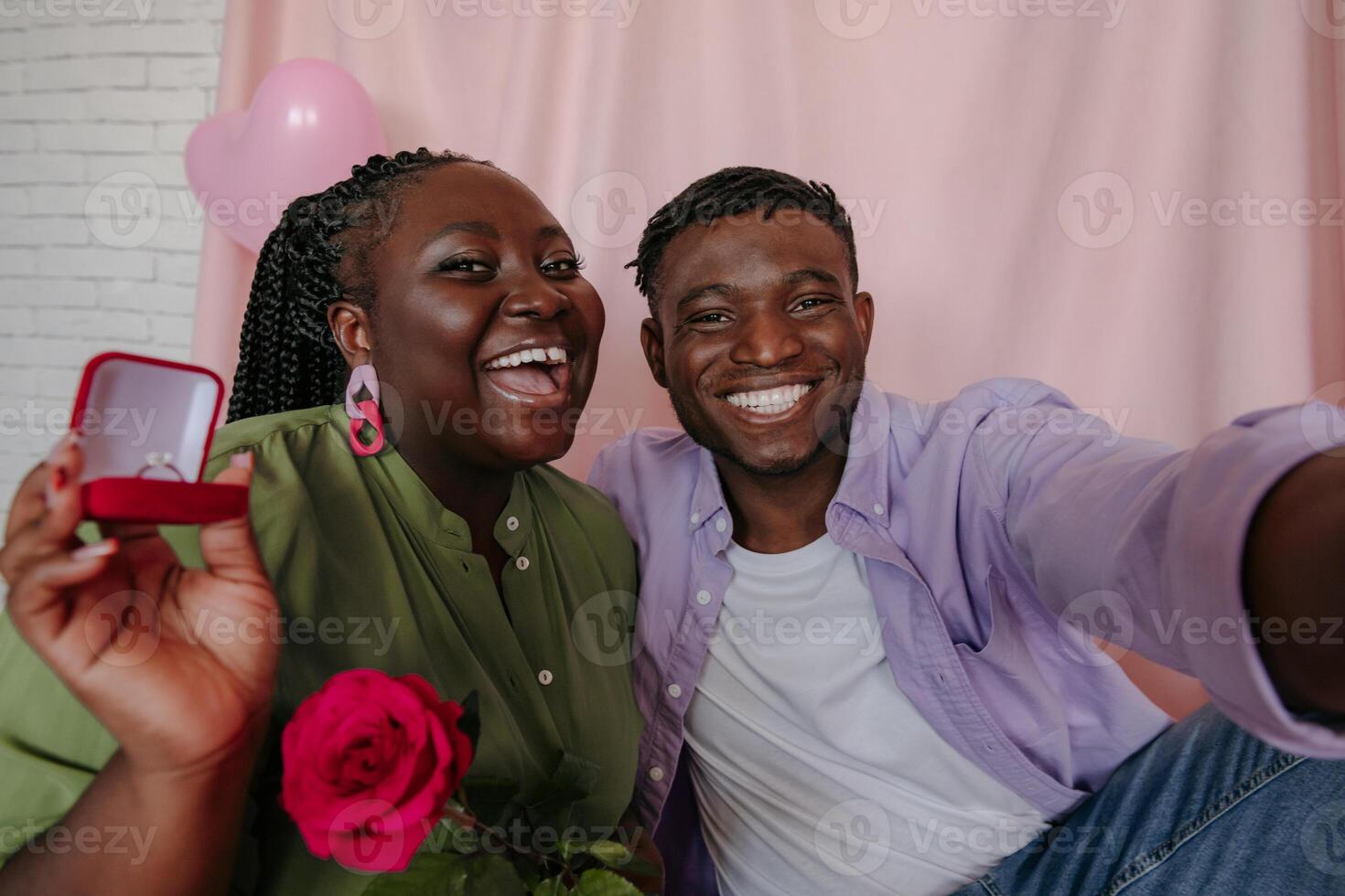 Happy young African woman holding engagement ring while embracing with her boyfriend on pink background photo