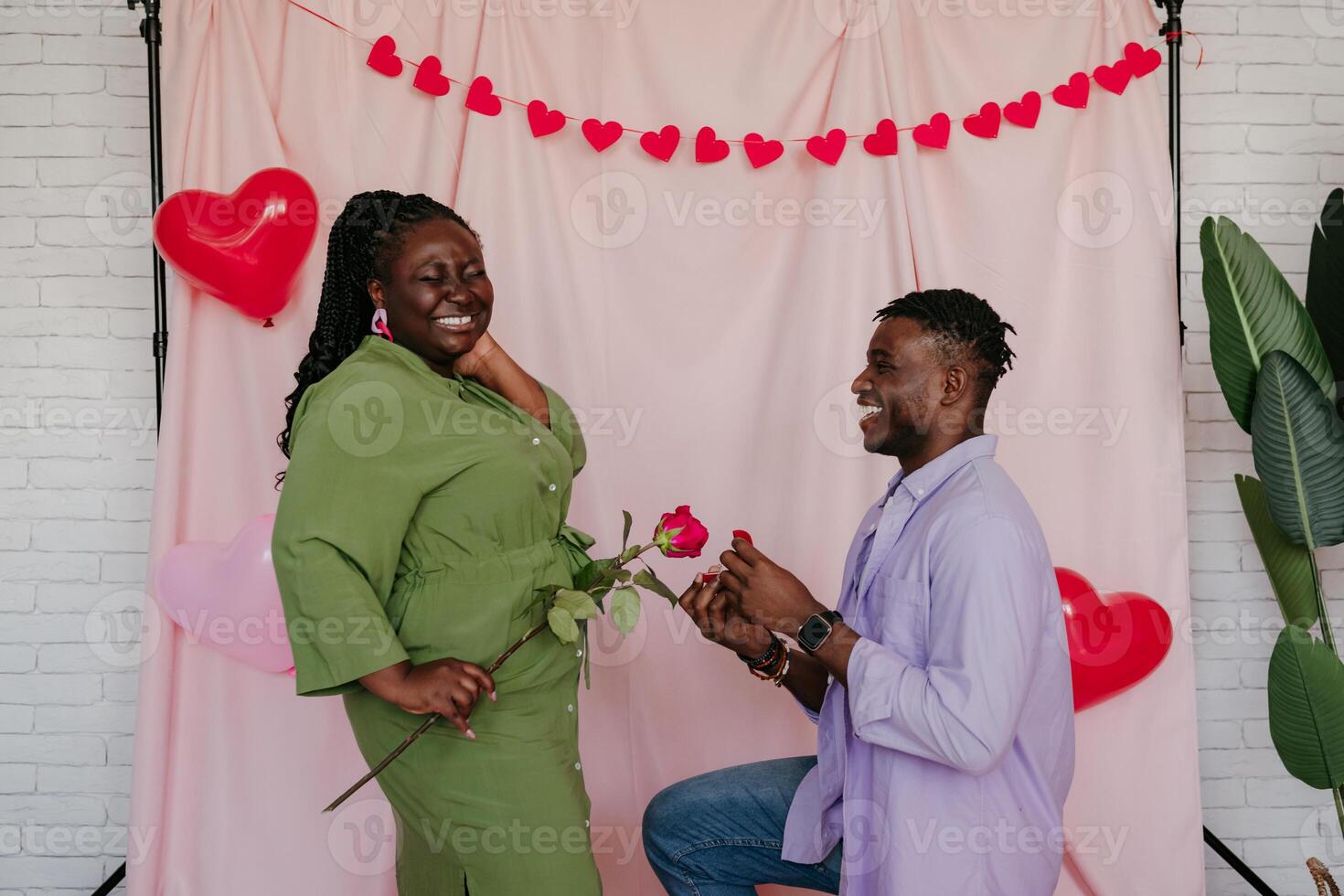 Handsome African man giving engagement ring to his surprised girlfriend while making proposal on pink background photo