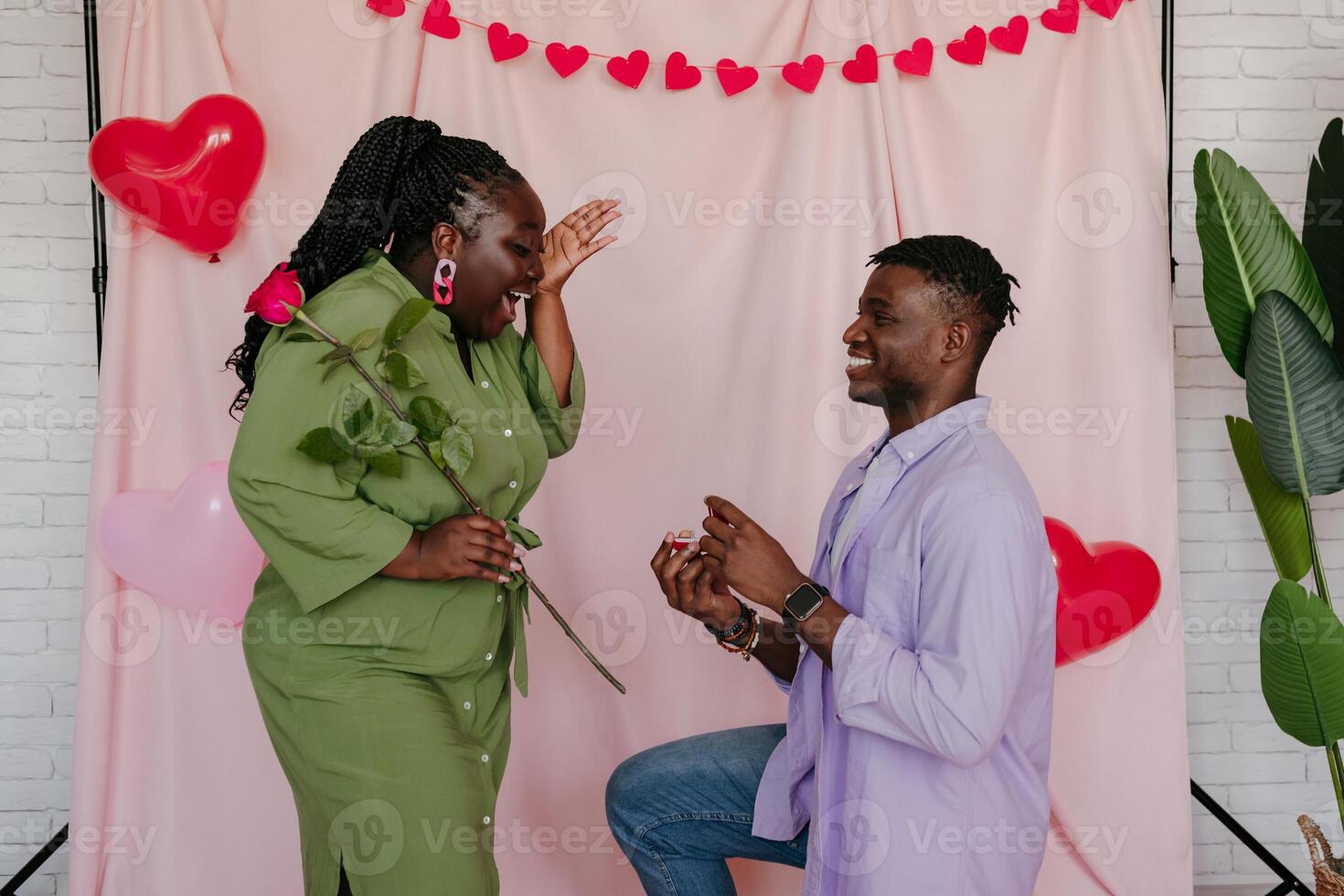 Young African man giving engagement ring to his surprised girlfriend while making proposal on pink background photo