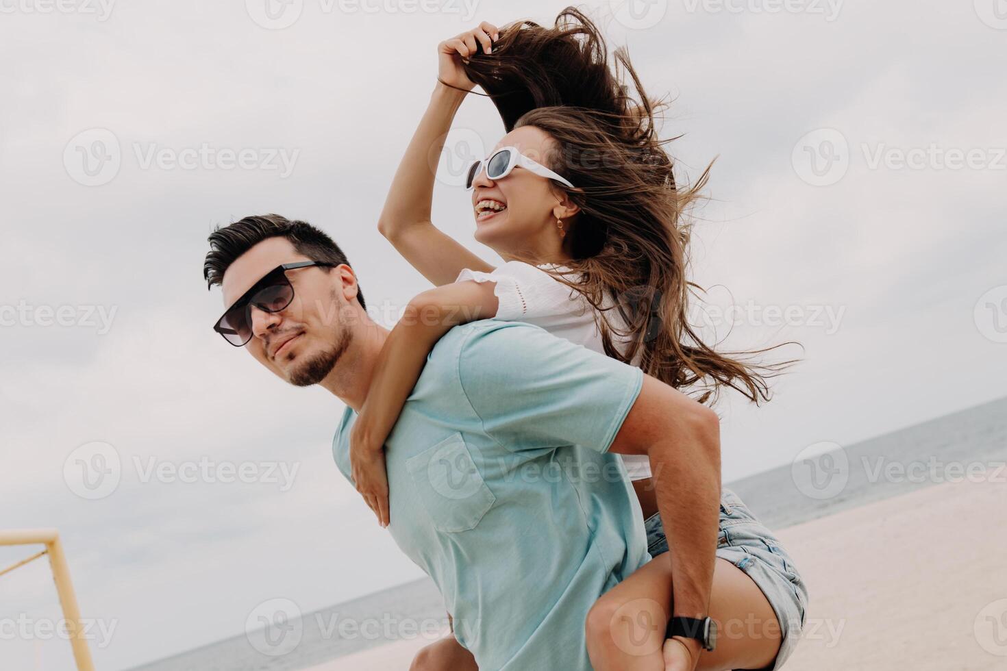 Handsome young man piggybacking his happy girlfriend while having fun on the beach together photo
