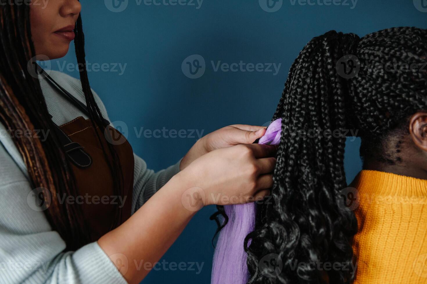 Close-up of African hairdresser braiding hair to female customer on blue background photo