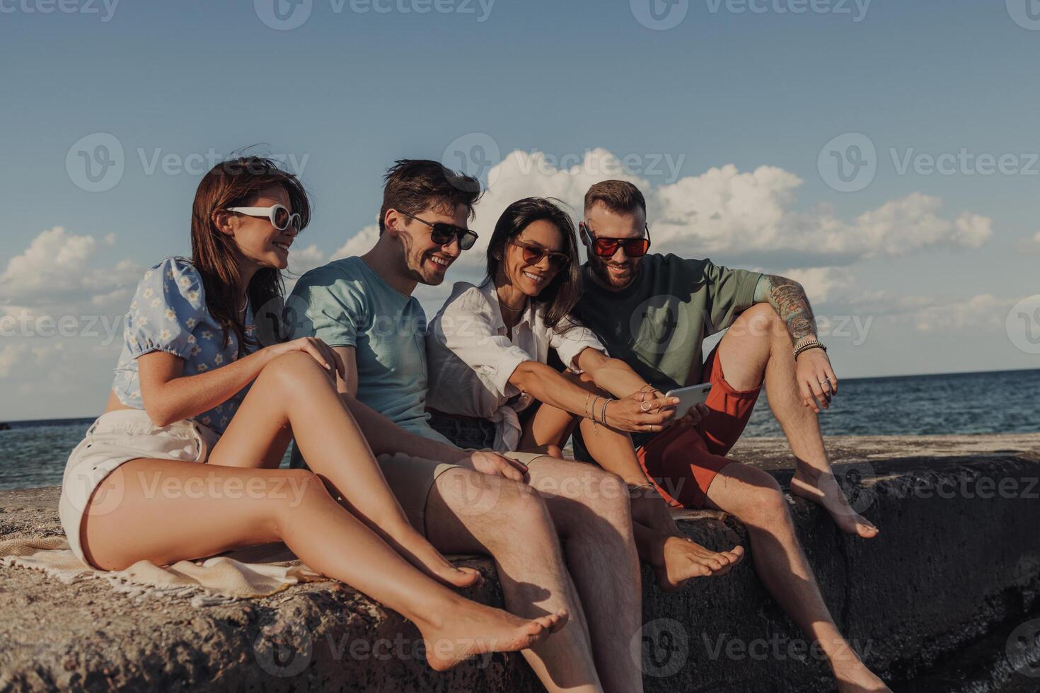 Group of young people looking at smart phone and smiling while enjoying time seaside together photo