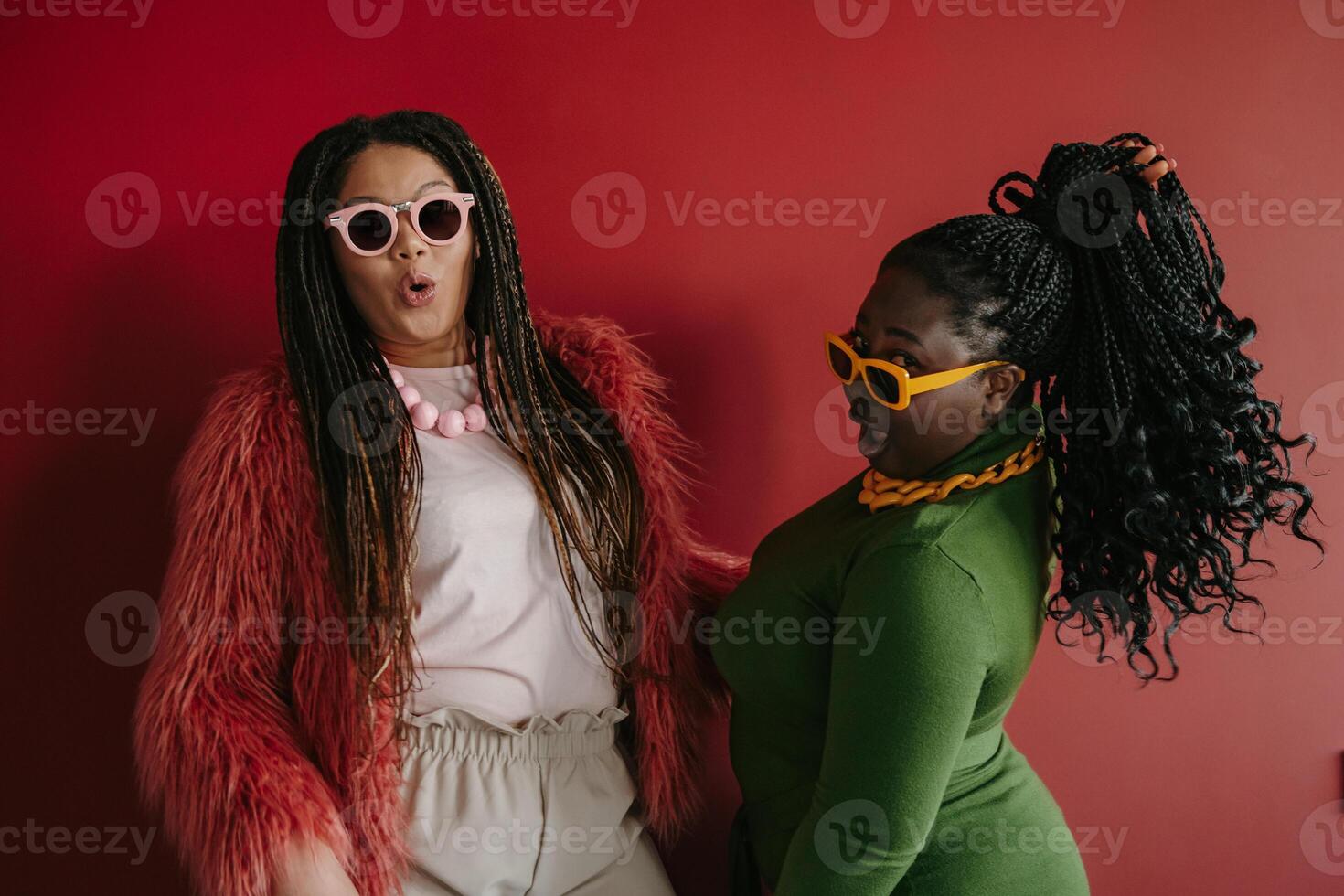Two playful plus size African woman with braided hair having fun while standing on red background together photo