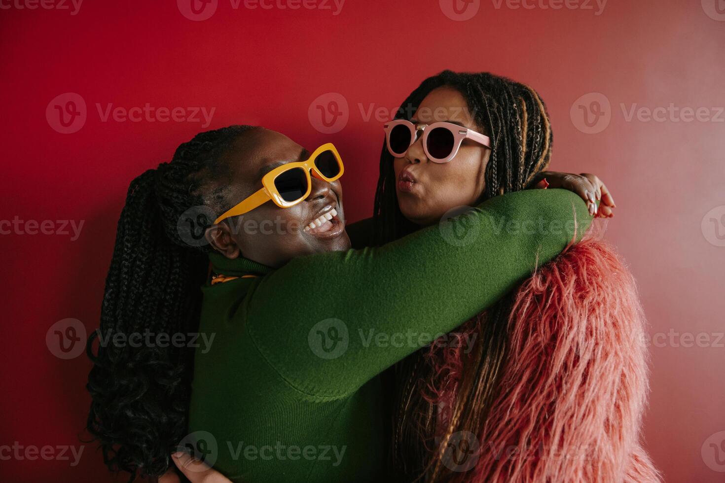 Two joyful plus size African woman with braided hair embracing on red background together photo