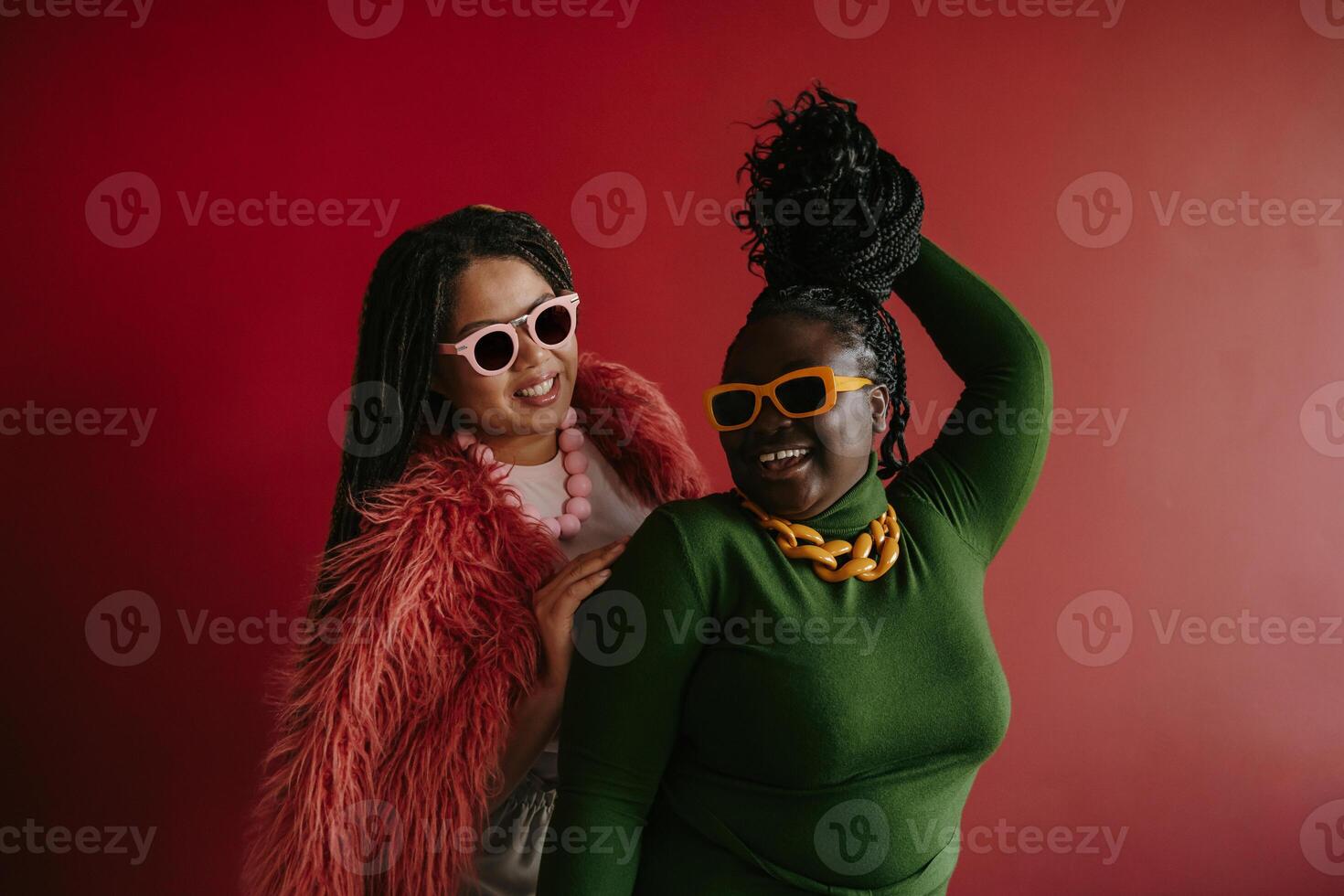 Two joyful plus size African woman playing with their braided hair while having fun on red background photo