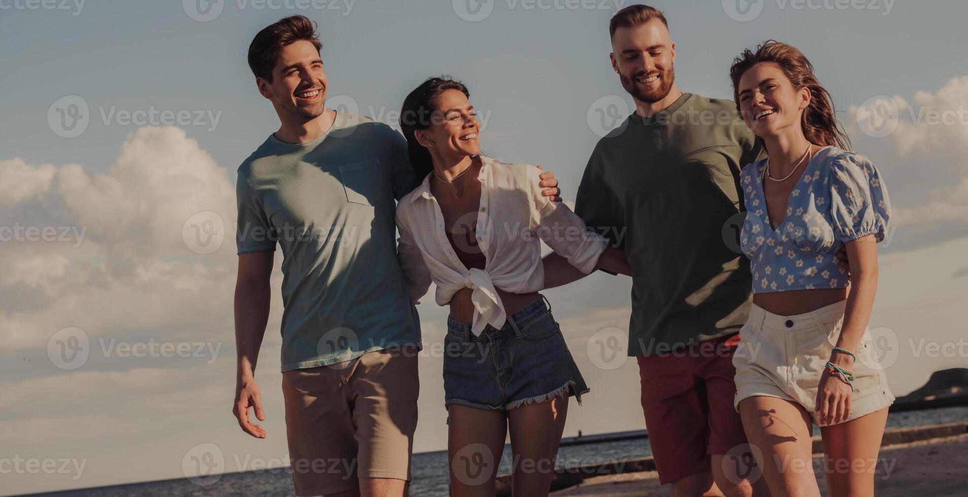 Group of cheerful young friends bonding and smiling while walking by the beach together photo