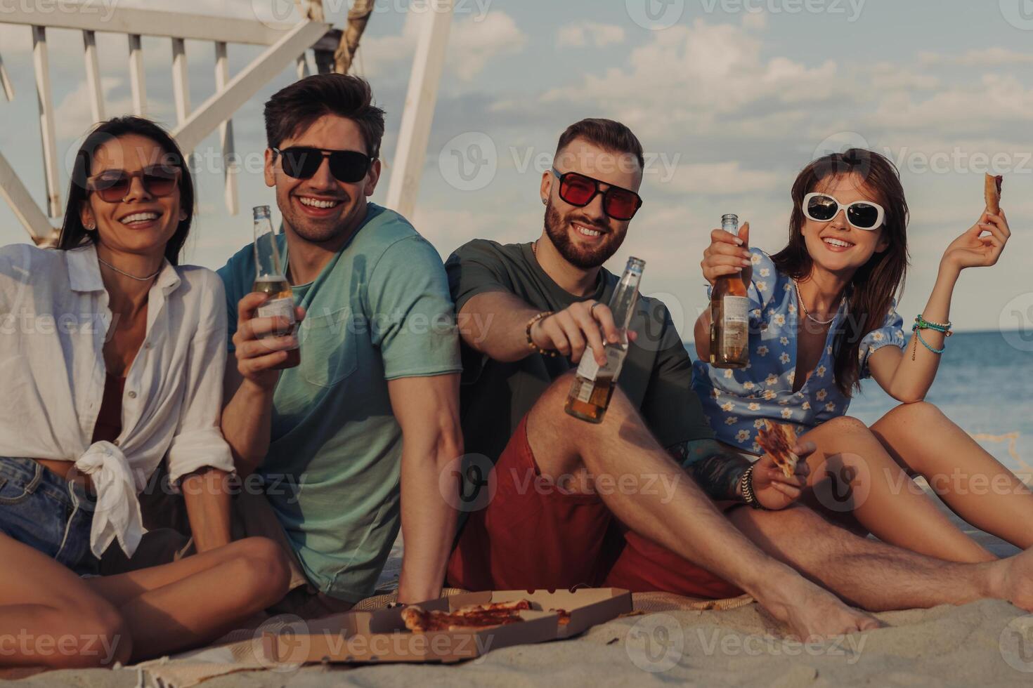 Four happy young friends looking at camera while enjoying pizza and beer on the beach photo