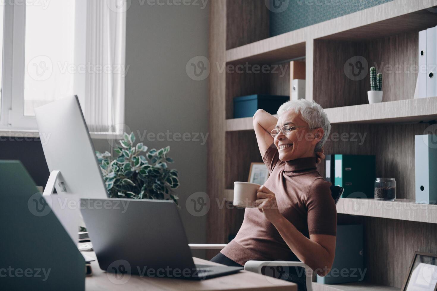 Relaxed senior businesswoman enjoying coffee and smiling while sitting at her working place photo