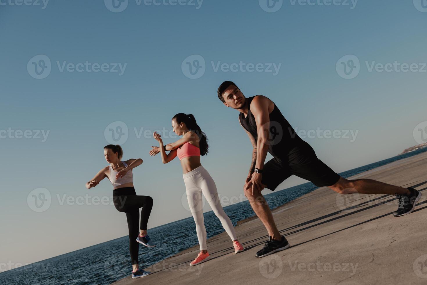 Three young people in sportswear doing stretching exercises before training seaside together photo
