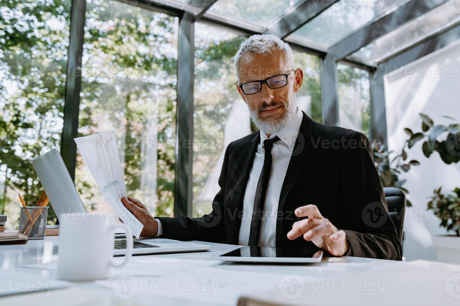 Confident mature businessman going through graphs and using technologies while working in office photo