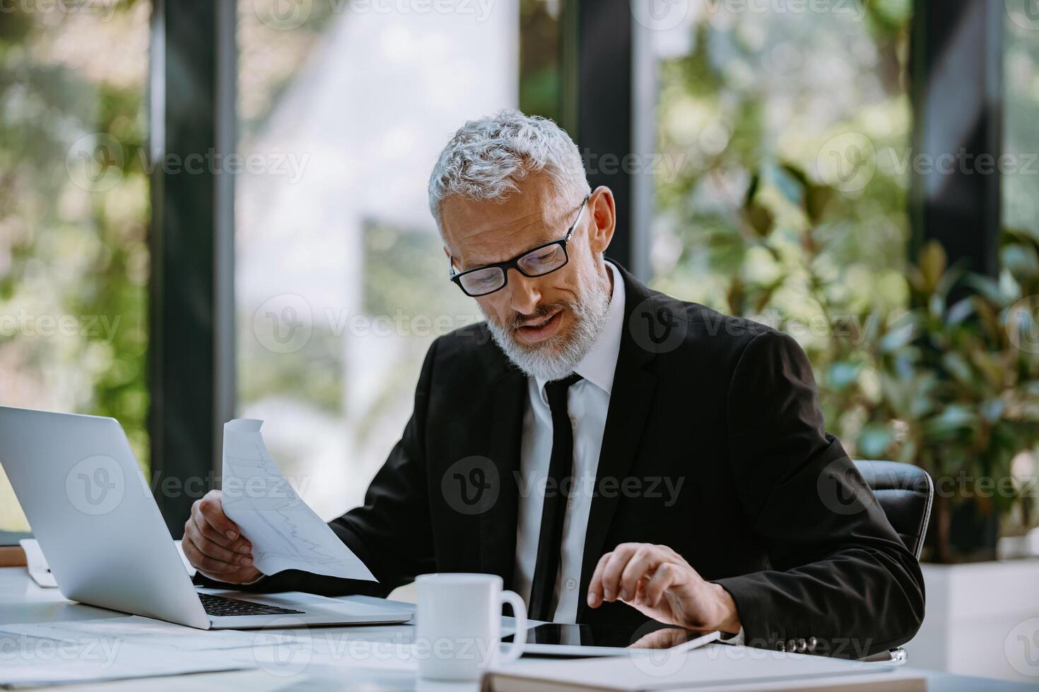 Confident mature businessman going through papers and using technologies while working in office photo