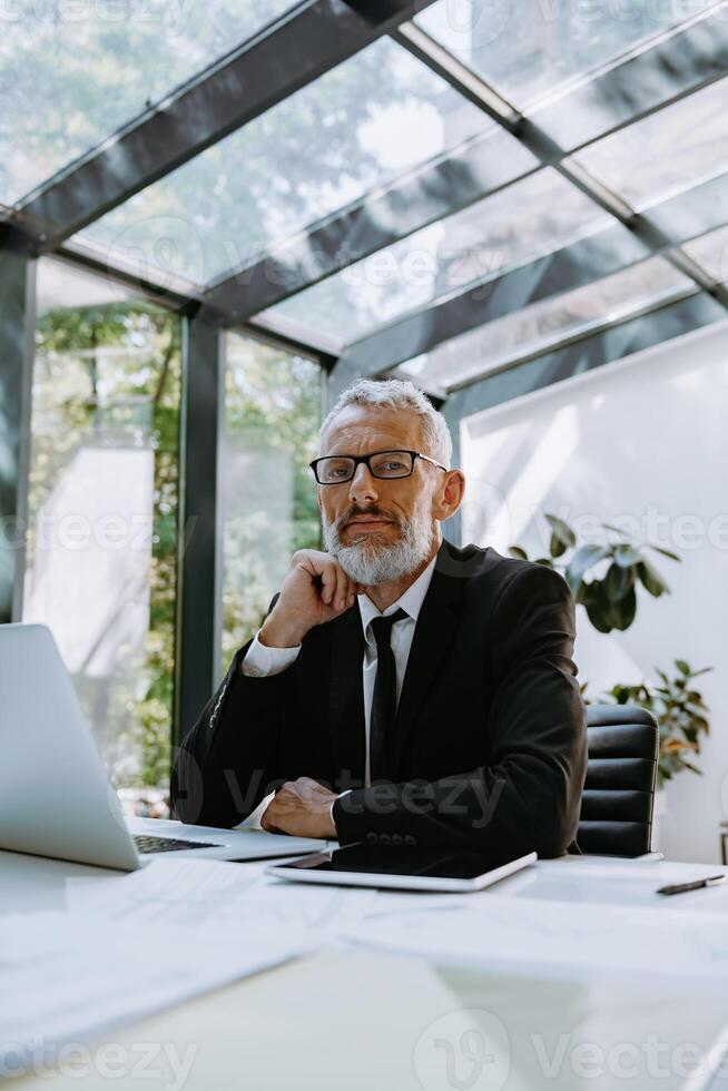 Confident mature businessman holding hand on chin while sitting at his working place in the office photo