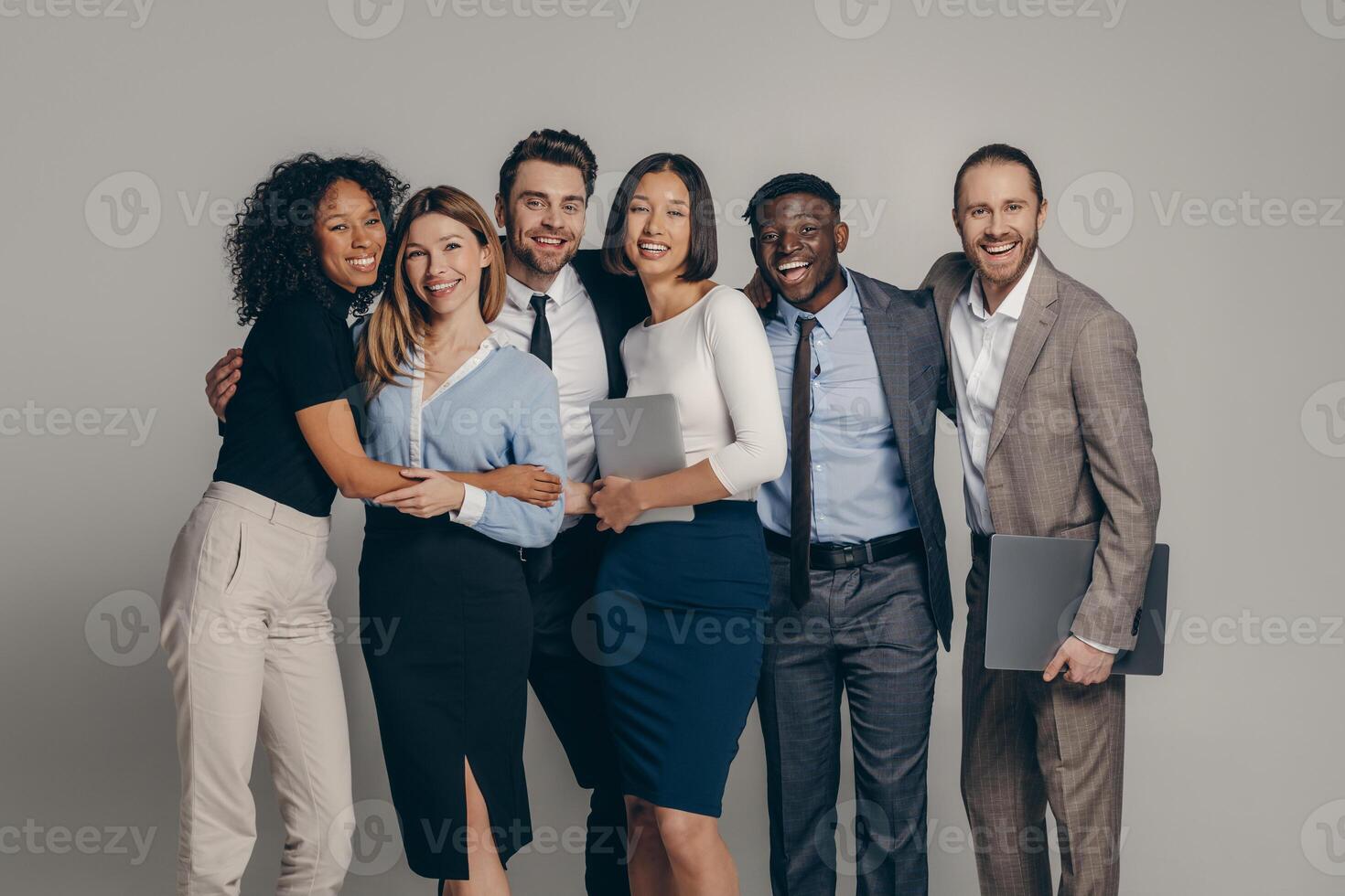 Smiling young business team in formalwear embracing while standing on beige background photo