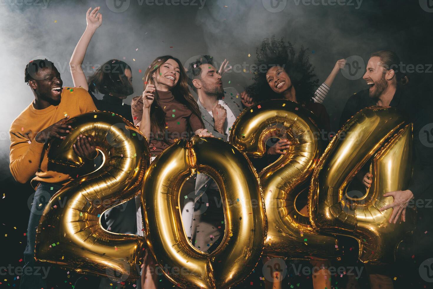 Group of joyful young people celebrating 2024 New Year while dancing and throwing confetti in night club photo