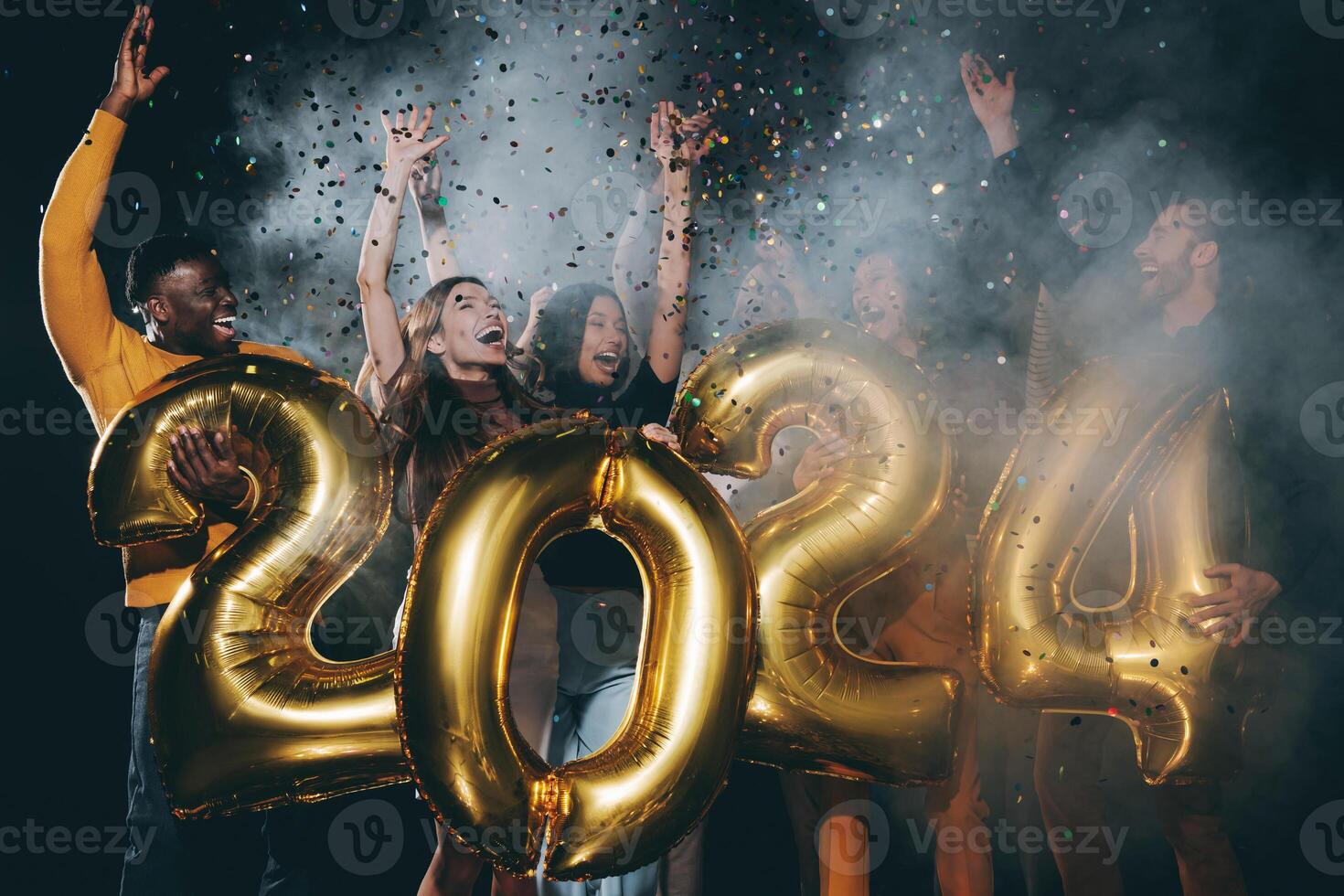 Group of joyful people holding 2024 numbers and throwing confetti while celebrating New Year in night club photo