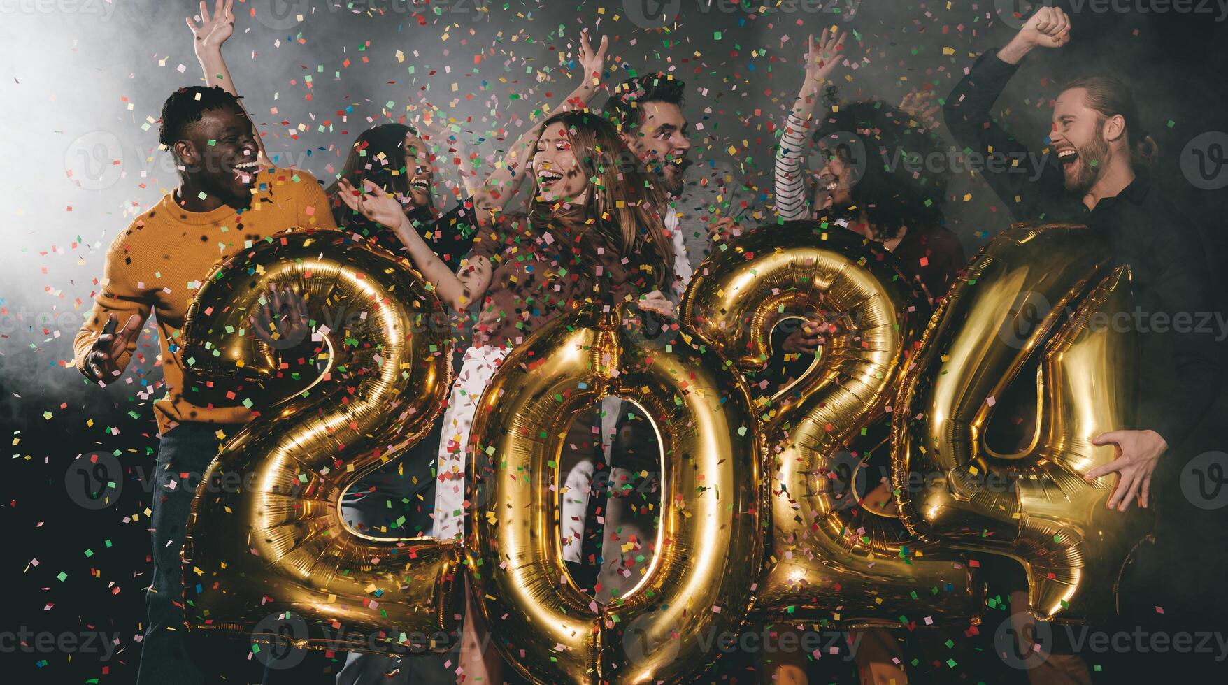 Group of happy people celebrating 2024 New Year while dancing and throwing confetti in night club photo
