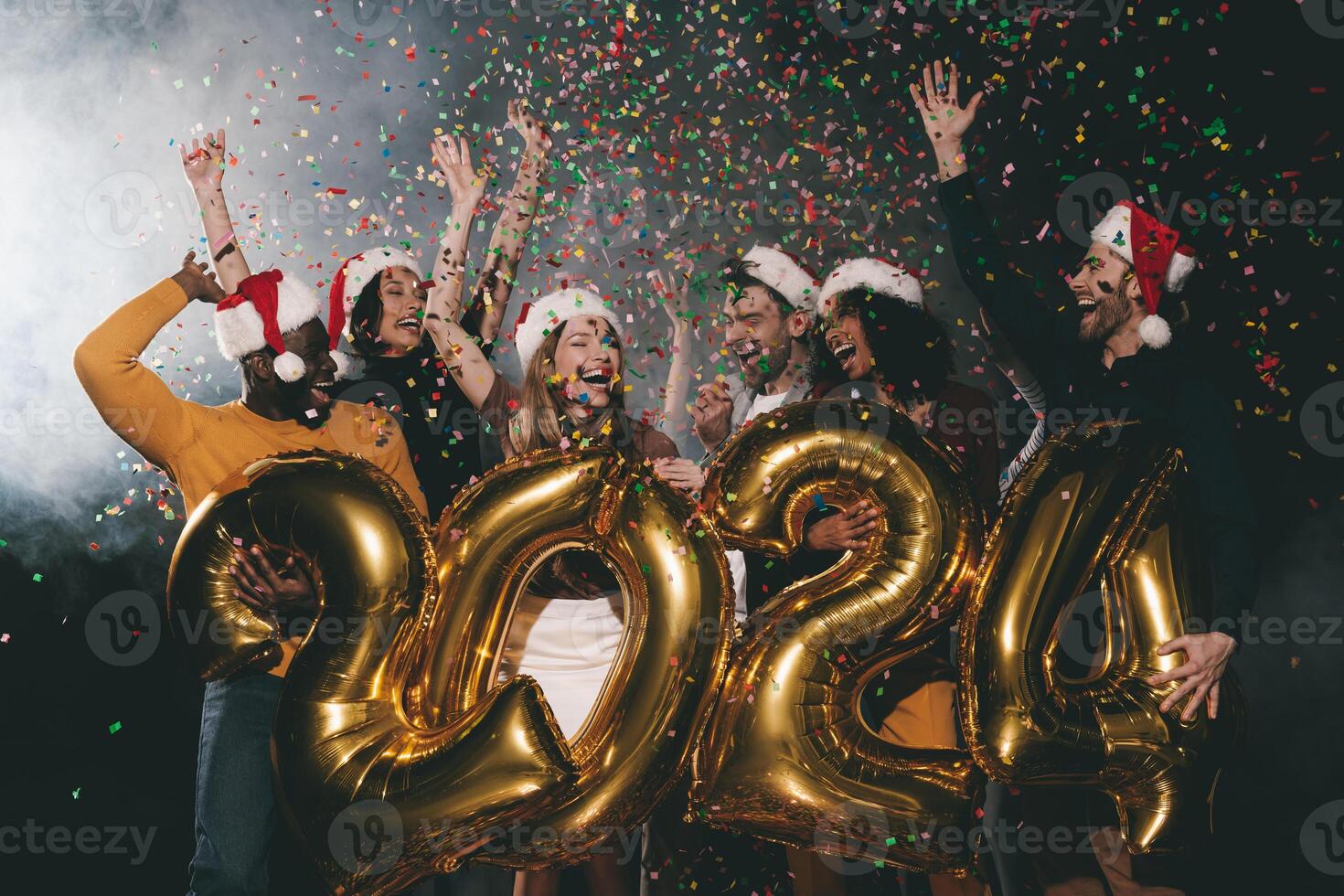 Group of joyful people in Christmas hats celebrating 2024 New Year while throwing confetti in night club photo
