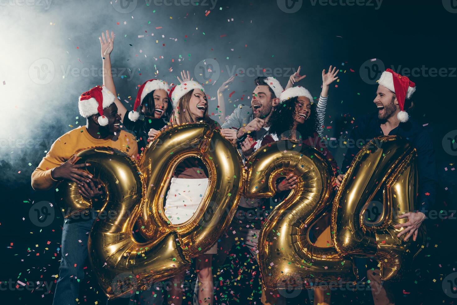 Group of happy people in Christmas hats celebrating 2024 New Year while throwing confetti in night club photo
