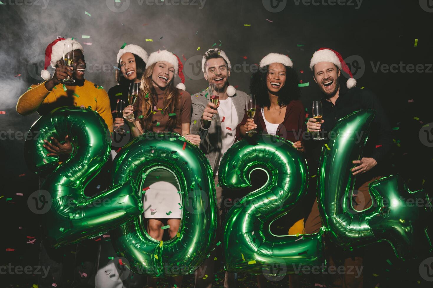 Group of joyful people in Christmas hats holding 2024 numbers while celebrating New Year in night club photo