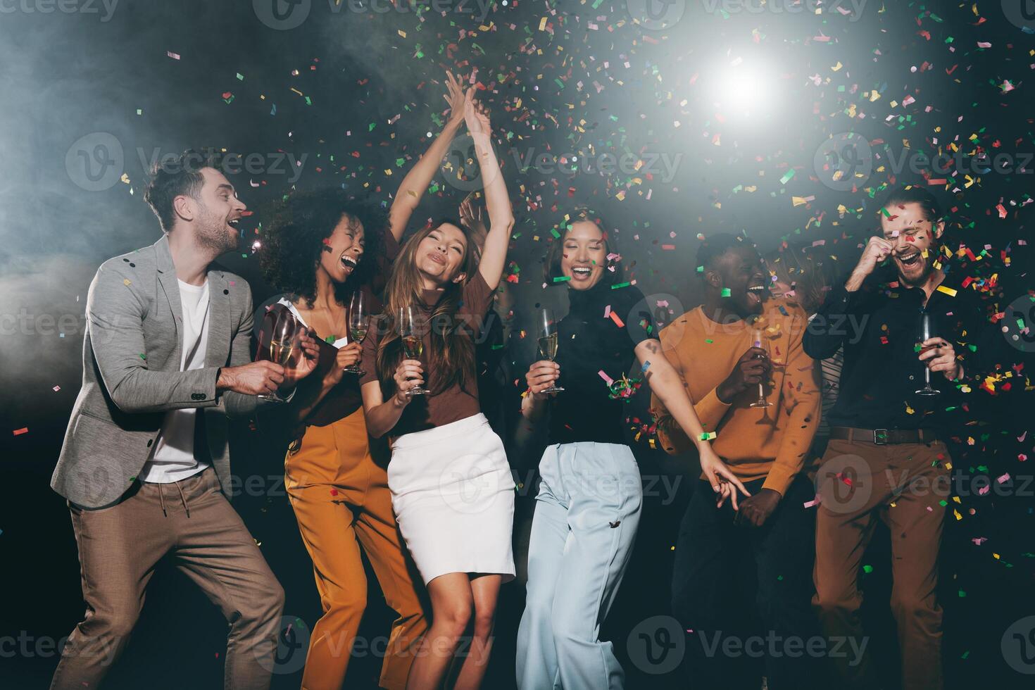 Group of young happy people enjoying champagne and throwing confetti while dancing in night club together photo