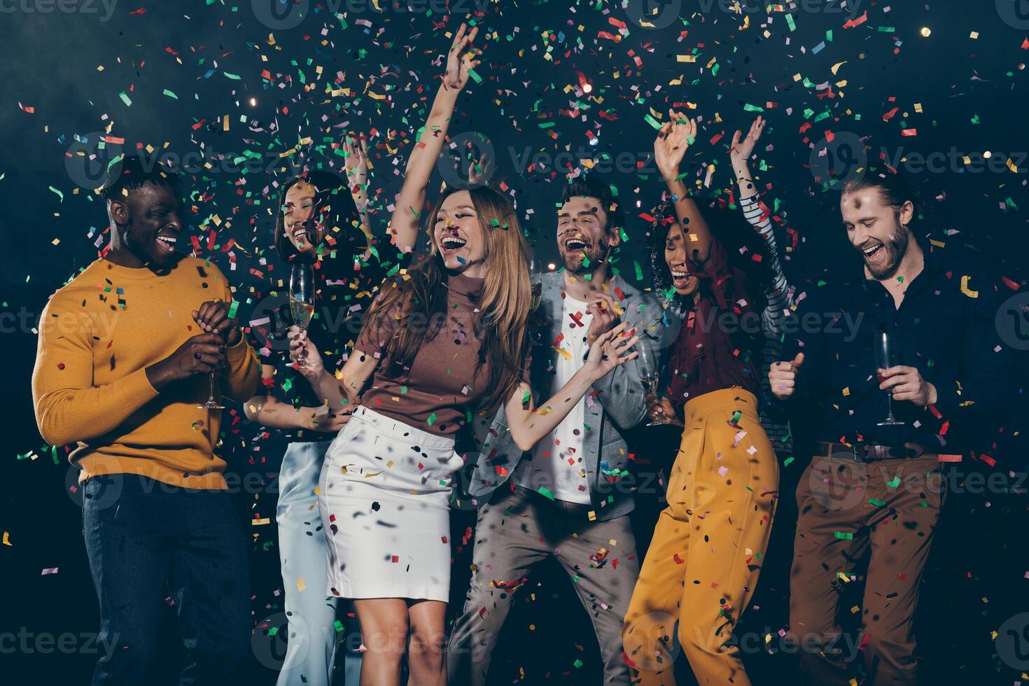 Group of young happy people enjoying champagne and throwing confetti while dancing in night club together photo