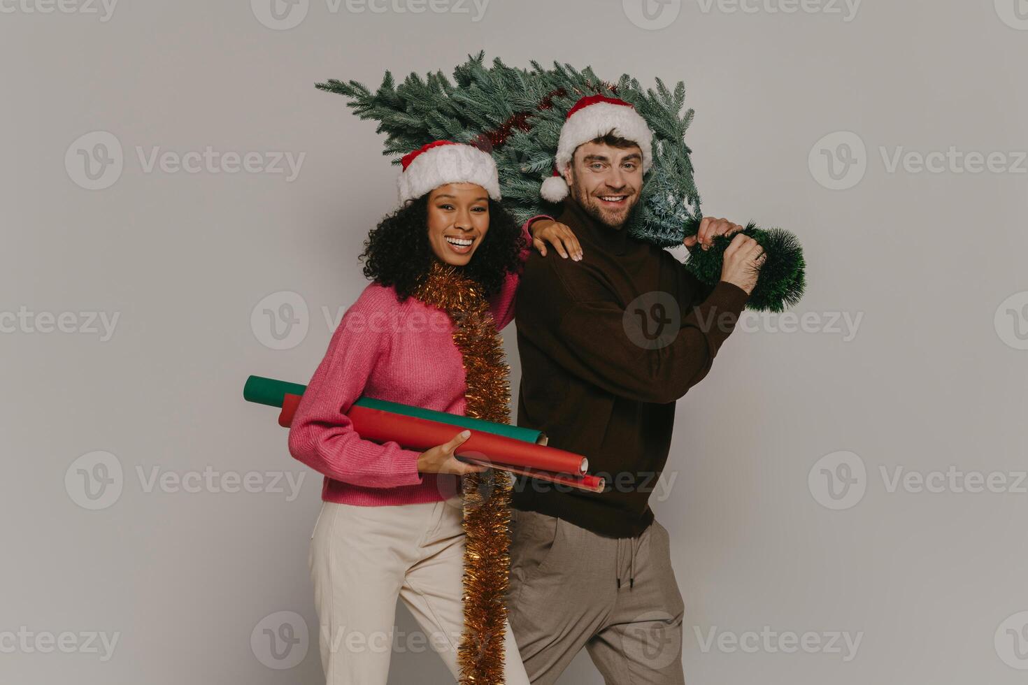 Happy young couple in Christmas hats bonding and smiling while man carrying Tree on shoulder photo
