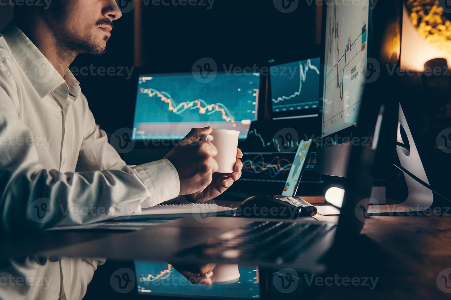 Male trader holding coffee cup while analyzing stock market data on multiple computers at night office photo