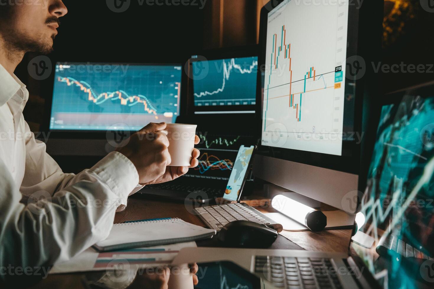 Male trader holding coffee cup while analyzing stock market data on multiple computers at night office photo