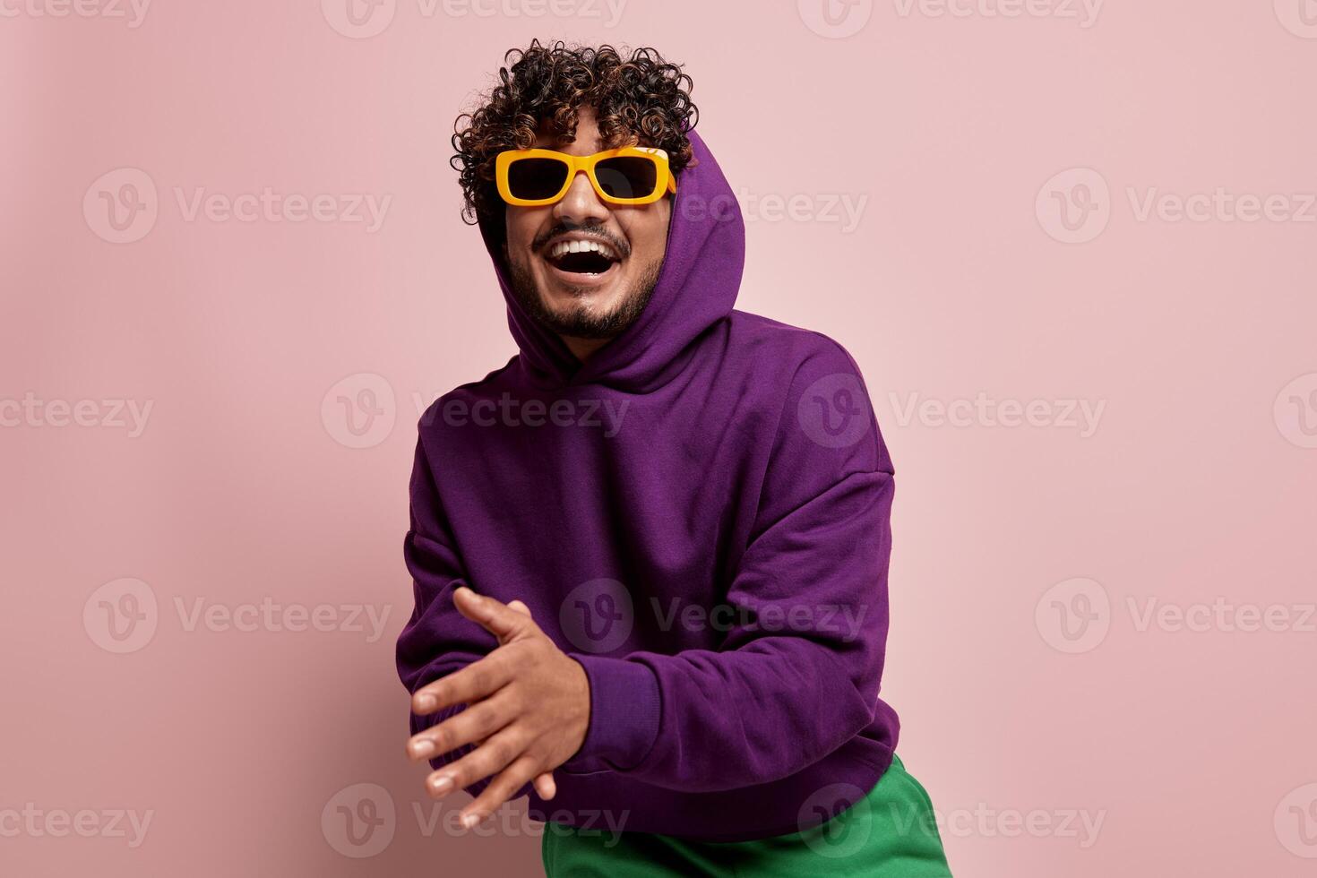 Happy young Indian man in stylish sunglasses smiling and gesturing against pink background photo