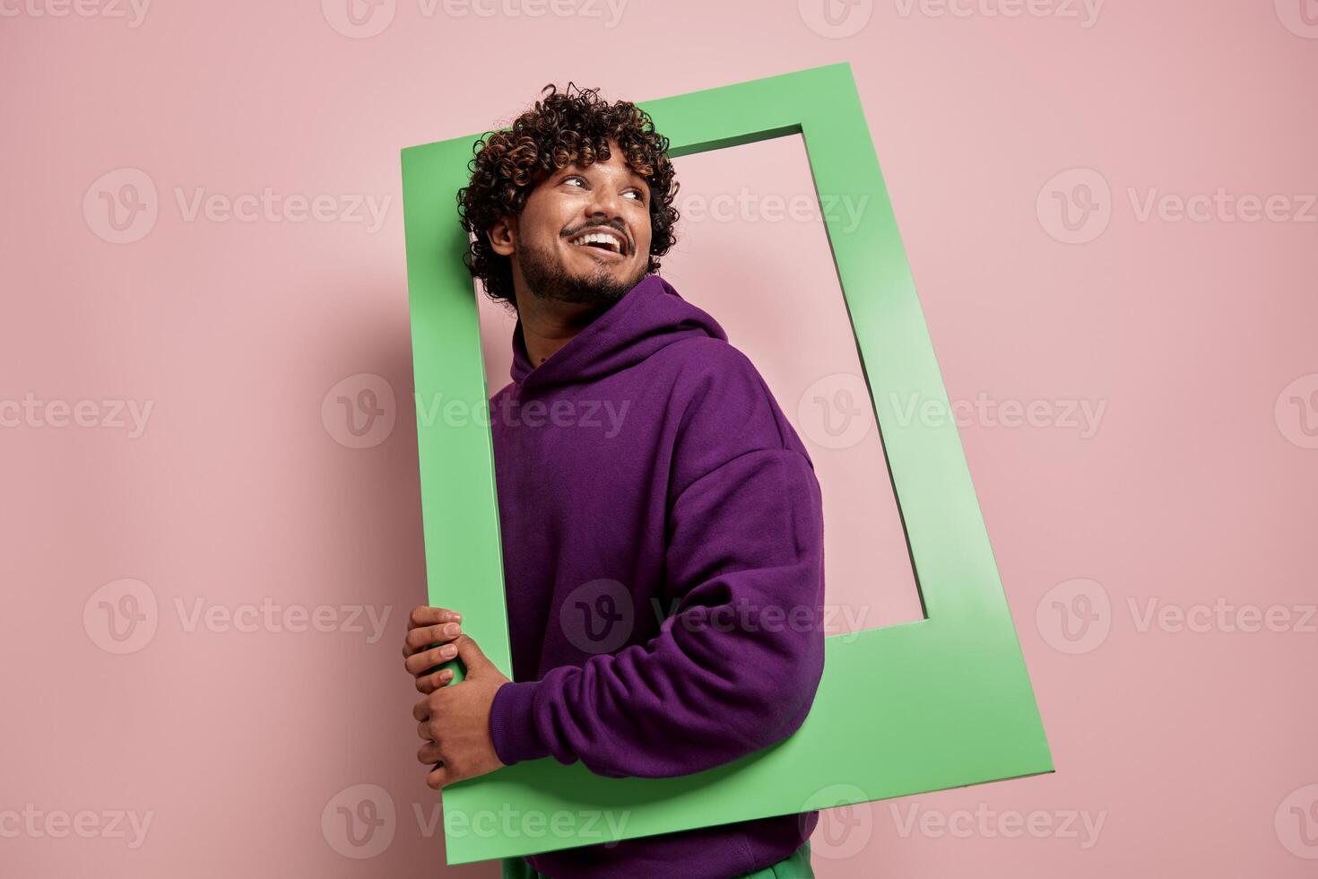 Joyful Indian man looking out of the picture frame while standing against pink background photo