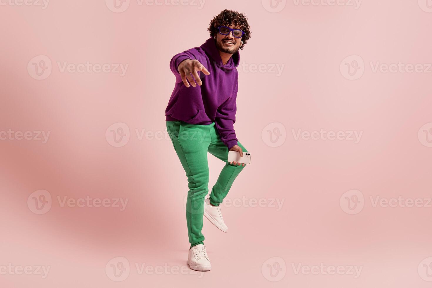 Young Indian man in headphones pointing camera and smiling against pink background photo