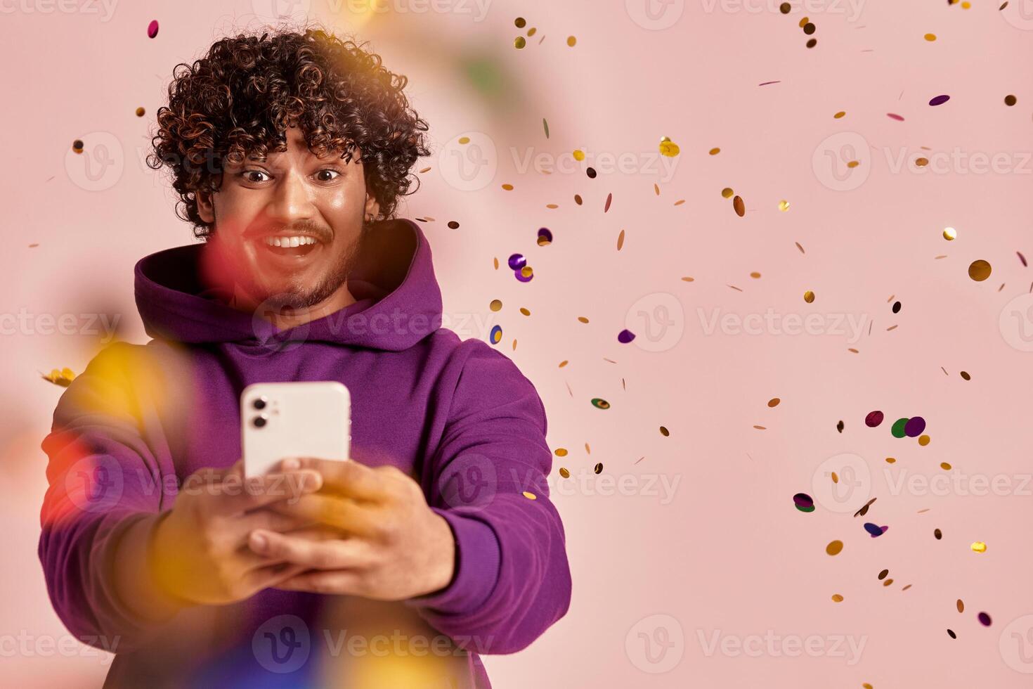 Joyful Indian man using smart phone while confetti flying around against pink background photo