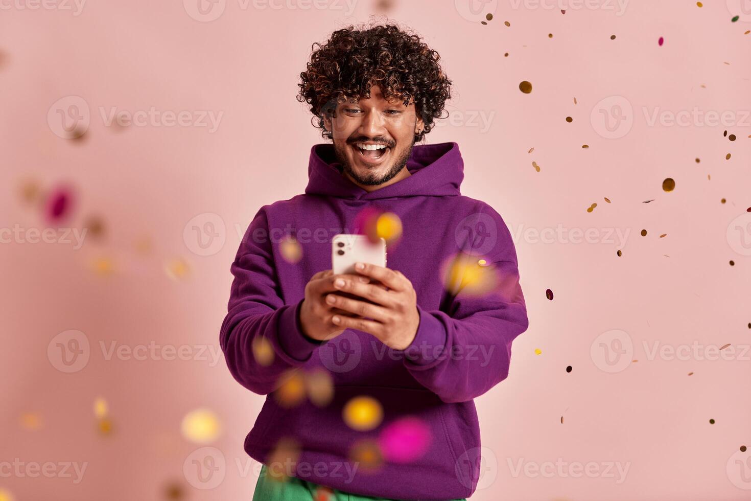 Excited Indian man using smart phone while confetti flying around against pink background photo
