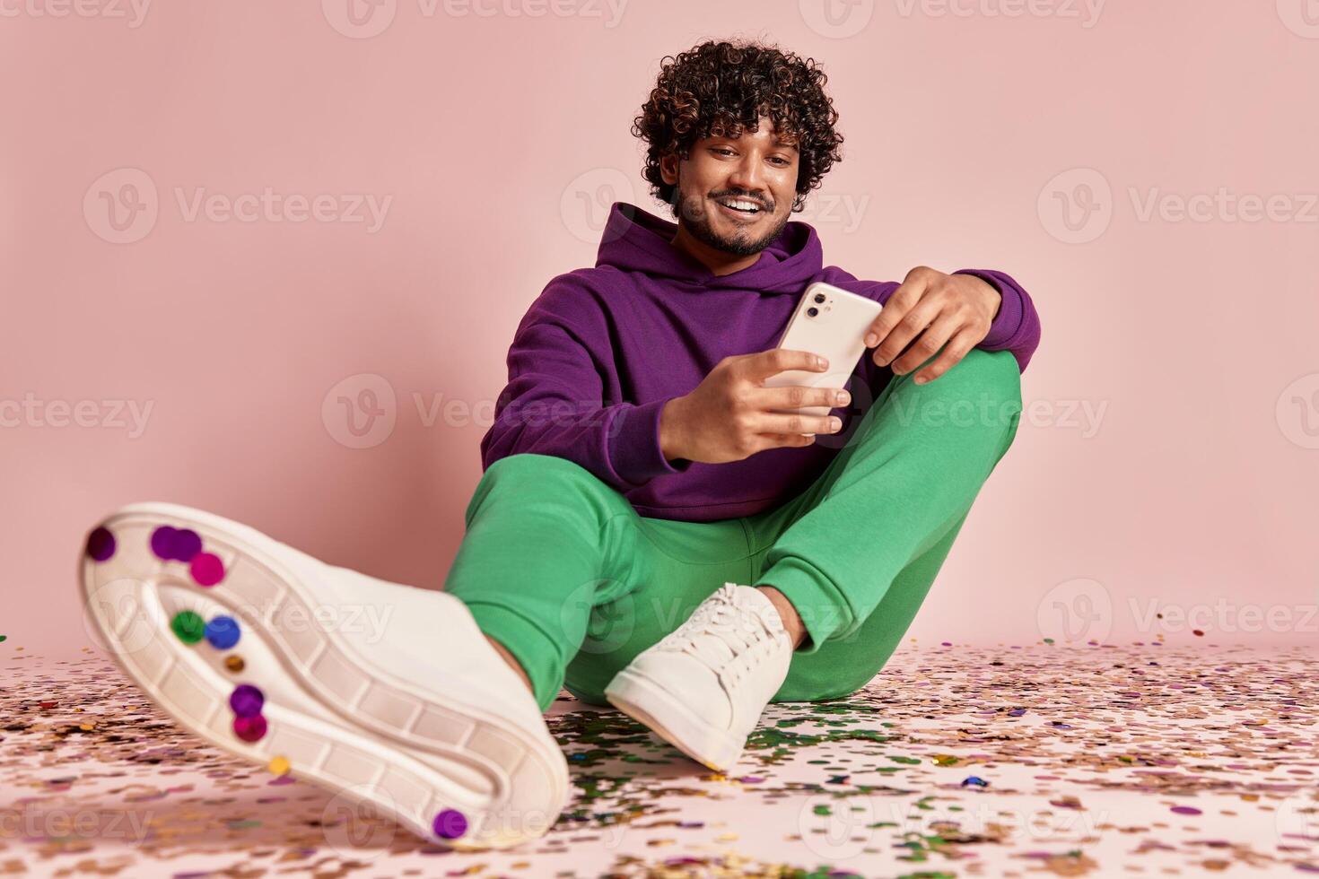Handsome Indian man using smart phone while sitting on confetti against pink background photo