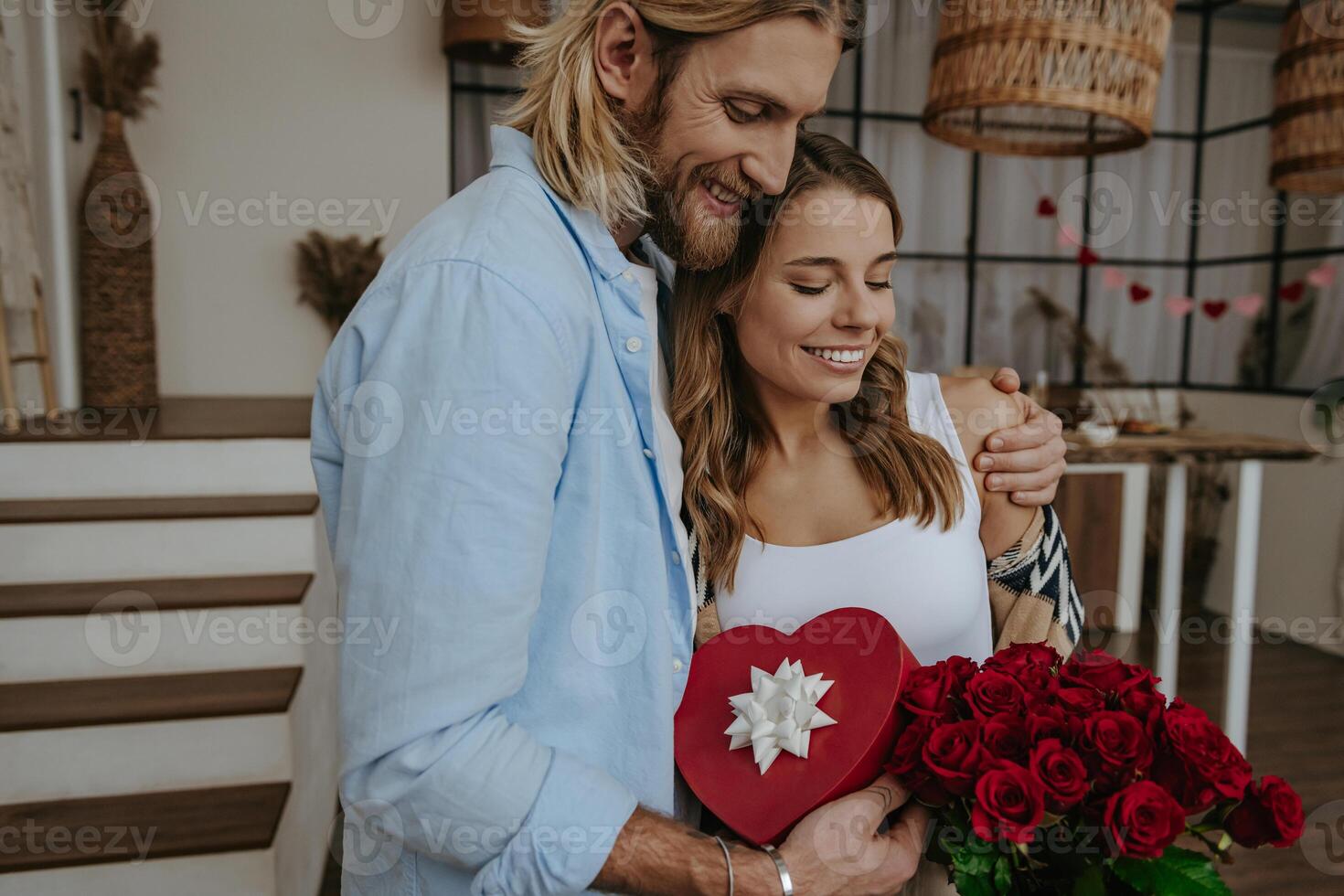 Young loving couple embracing while holding red roses and gift box with decorated living room on background photo