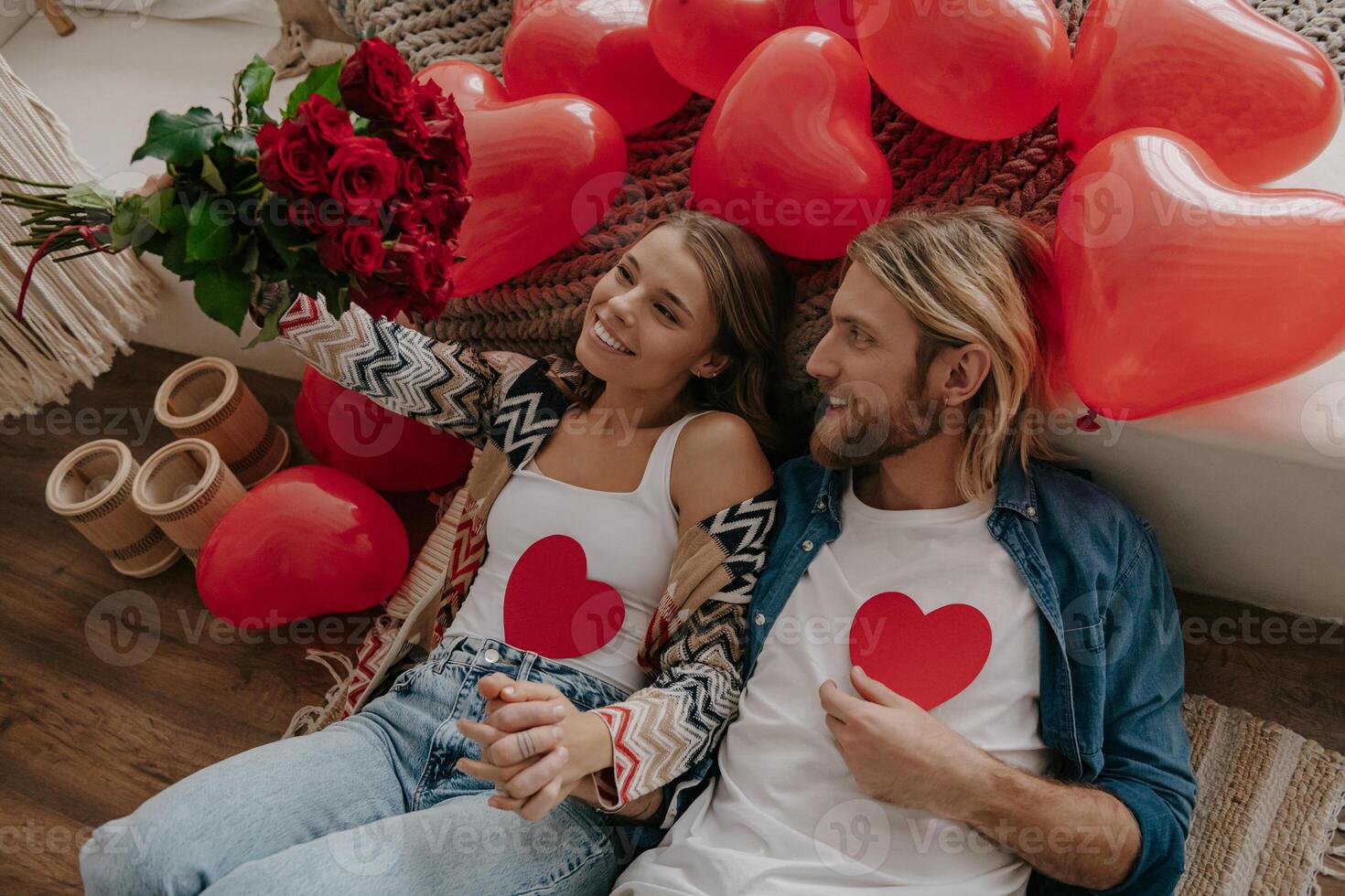 Top view of loving couple holding heart shape Valentines cards while leaning on bed surrounded with red balloons photo
