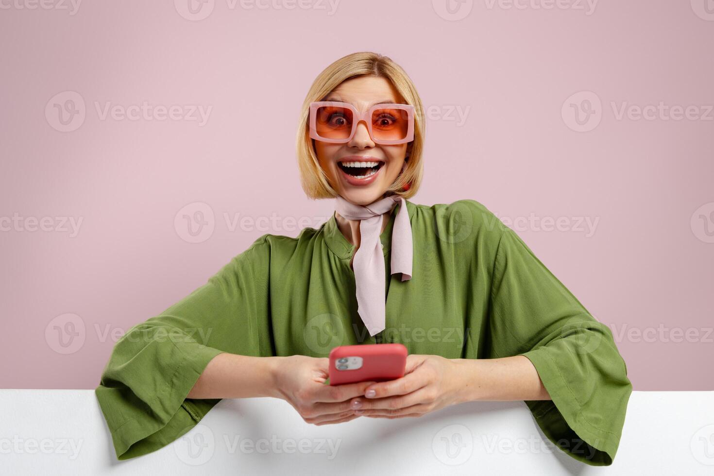 Fashionable young woman holding smart phone and looking excited against pink background photo
