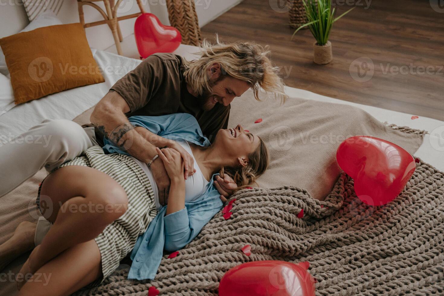 Playful loving couple embracing and having fun while sitting on bed surrounded with red heart shape balloons photo