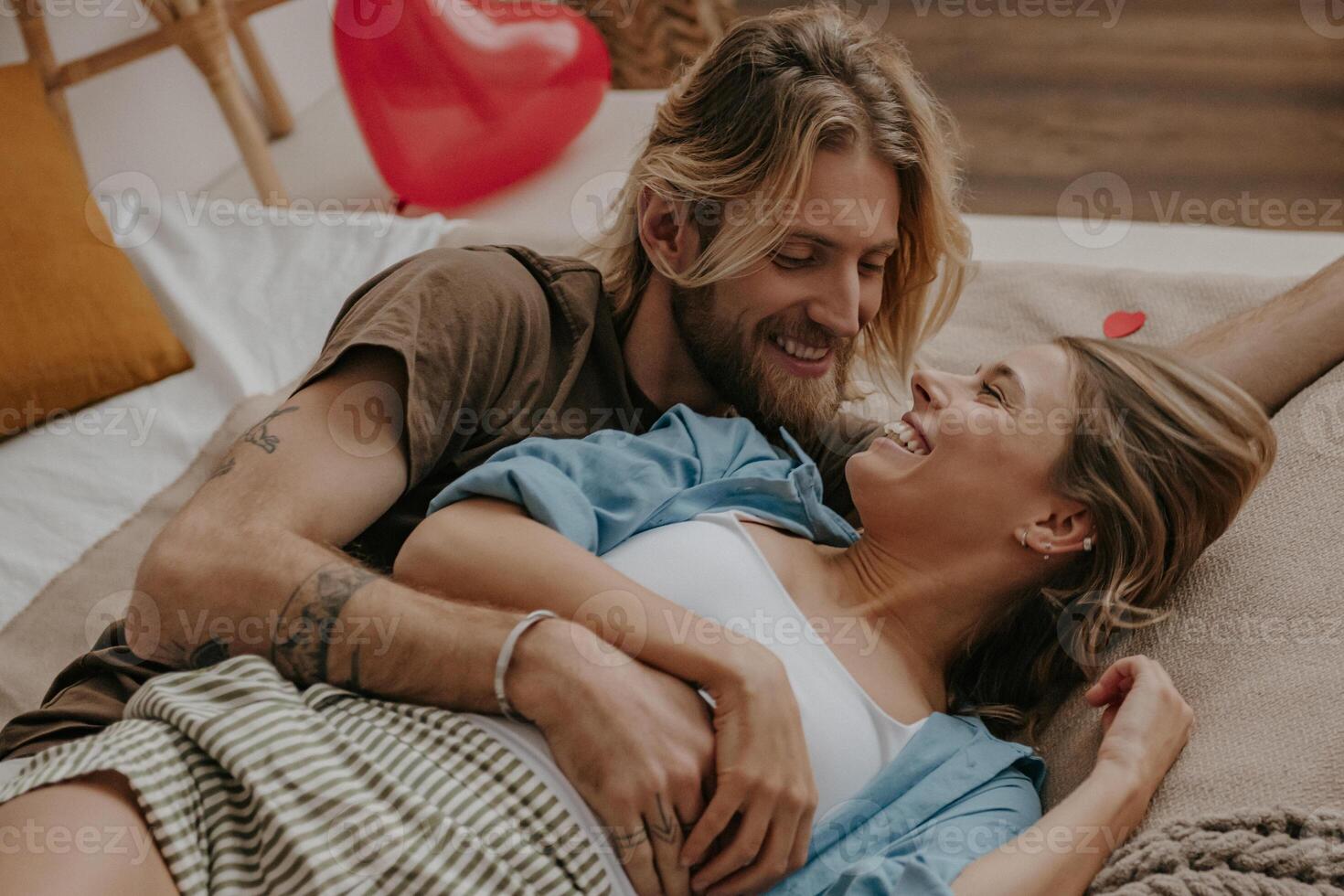Relaxed loving couple embracing and smiling while lying in bed surrounded with red heart shape balloons photo