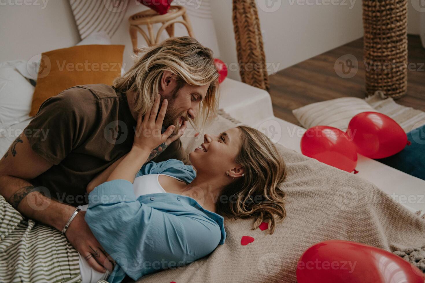 Relaxed loving couple embracing and smiling while lying in bed surrounded with red heart shape balloons photo