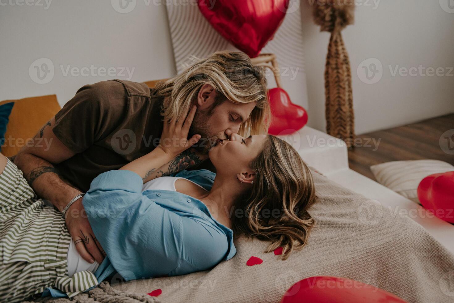 Passionate loving couple embracing and kissing while lying down in bed surrounded with red heart shape balloons photo