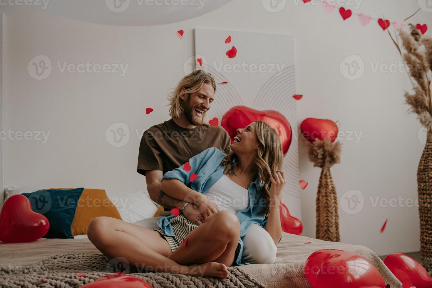 Joyful loving couple sitting on bed and throwing heart shape confetti while celebrating Valentines day at home photo