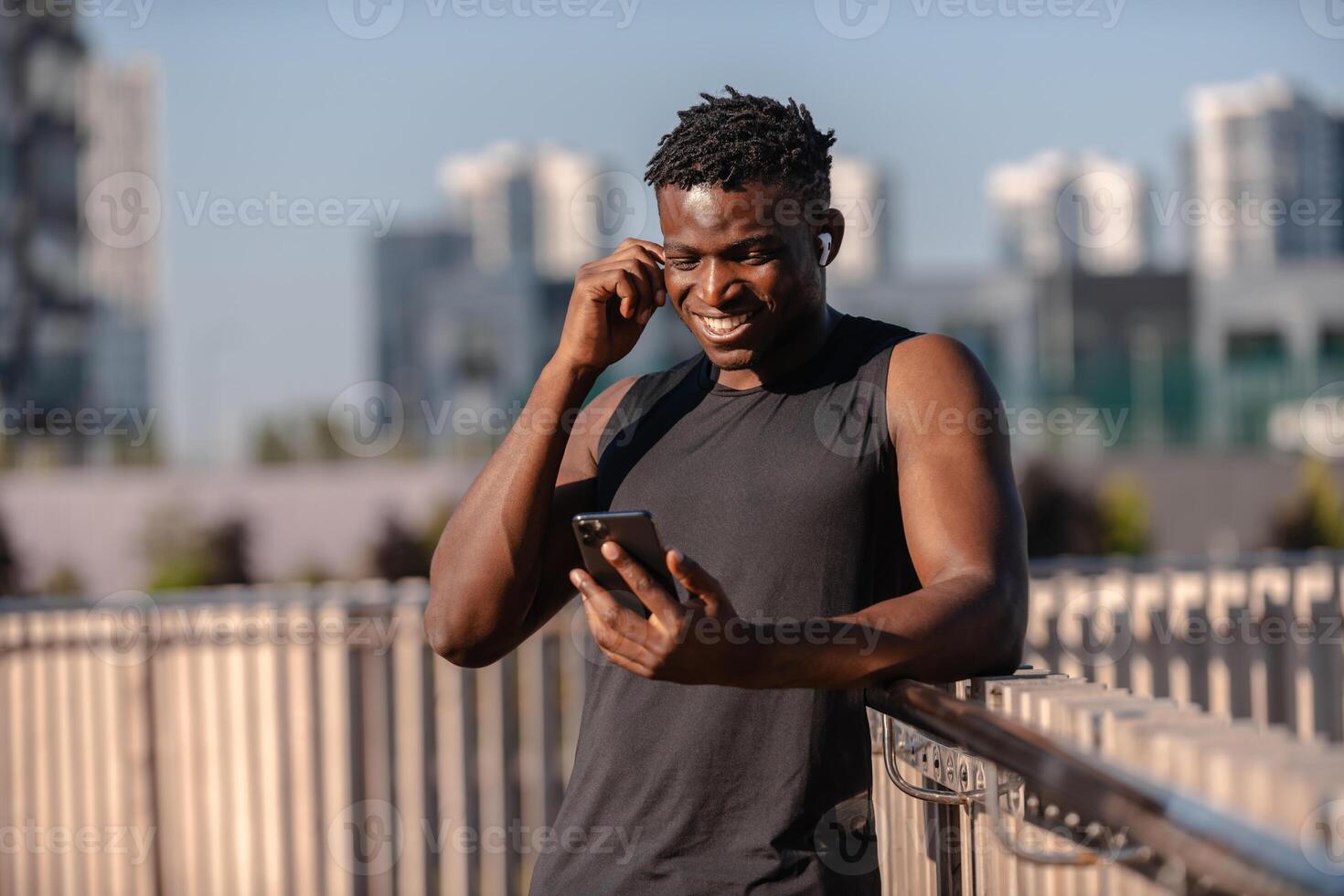 Cheerful African man in sportswear using smart phone and adjusting headphones while resting after training photo