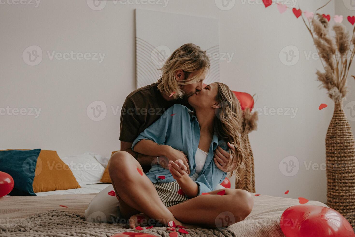 Romantic loving couple sitting on bed and kissing while red heart shape confetti flying around photo